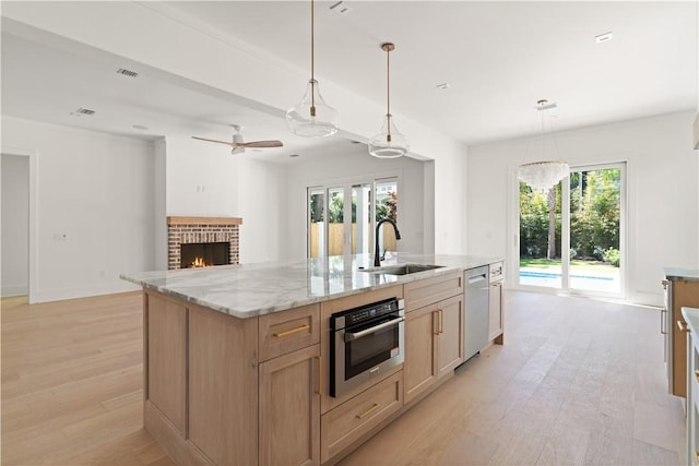 kitchen with a brick fireplace, a center island with sink, light wood-type flooring, a sink, and appliances with stainless steel finishes