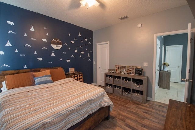 bedroom featuring visible vents, an accent wall, a textured ceiling, wood finished floors, and baseboards