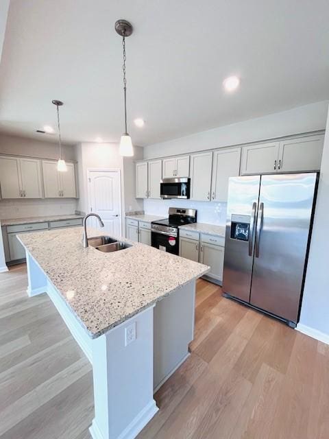 kitchen with light wood-type flooring, an island with sink, a sink, light stone counters, and stainless steel appliances