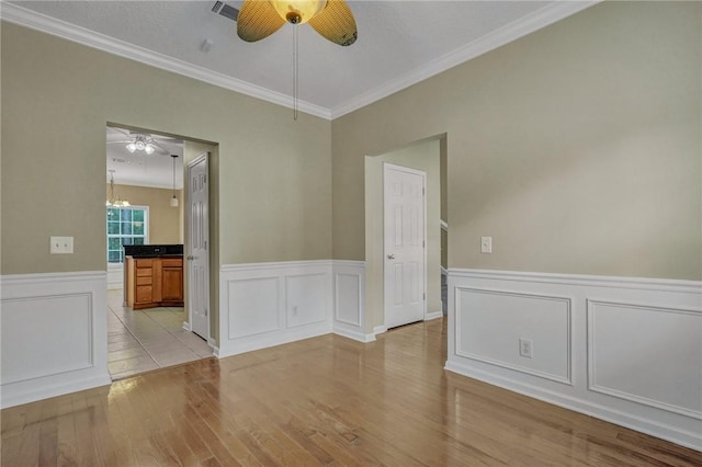 empty room with wainscoting, light wood-style flooring, ornamental molding, and ceiling fan with notable chandelier