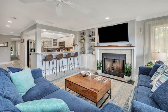 tiled living room featuring ceiling fan, ornamental molding, a tile fireplace, and built in shelves