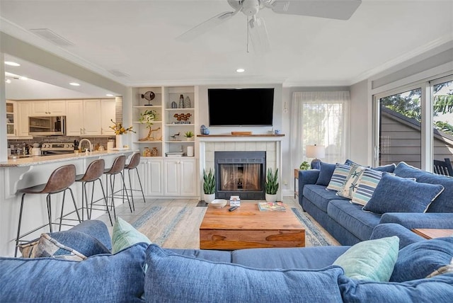 living room featuring ceiling fan, sink, light hardwood / wood-style flooring, a tiled fireplace, and ornamental molding