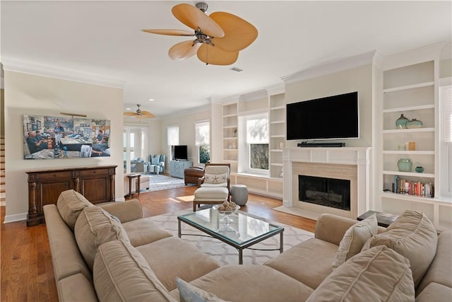 living room with ceiling fan, built in features, light wood-type flooring, a fireplace, and ornamental molding