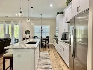 kitchen with sink, hanging light fixtures, a center island with sink, stainless steel fridge, and white cabinets