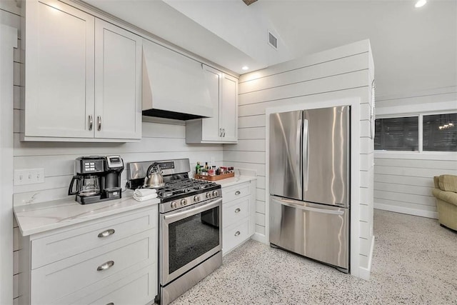 kitchen featuring white cabinetry, stainless steel appliances, light stone counters, custom exhaust hood, and wood walls