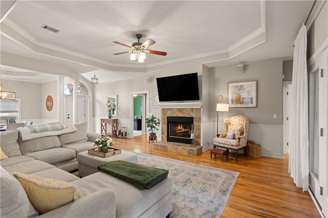 living room with ornamental molding, a tray ceiling, ceiling fan, a tiled fireplace, and light hardwood / wood-style floors