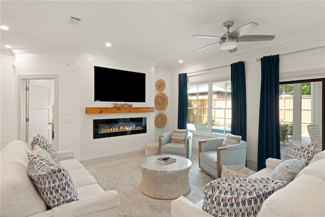 living area with ceiling fan, a glass covered fireplace, crown molding, visible vents, and plenty of natural light