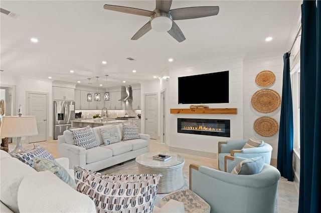 living area featuring visible vents, a glass covered fireplace, recessed lighting, ornamental molding, and a ceiling fan