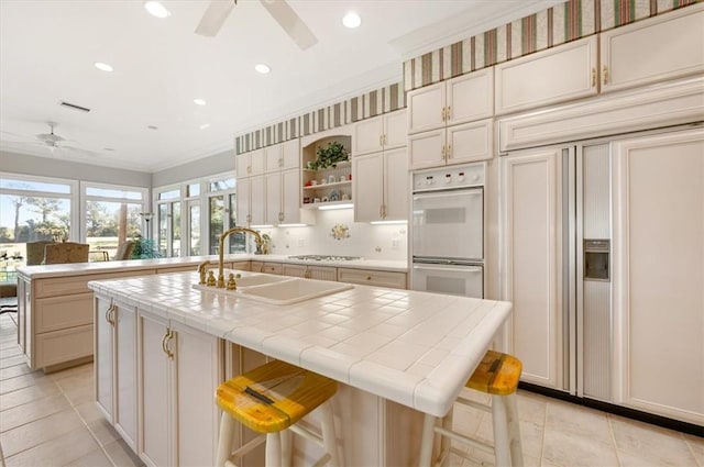 kitchen featuring double oven, sink, a center island with sink, tile counters, and a breakfast bar area