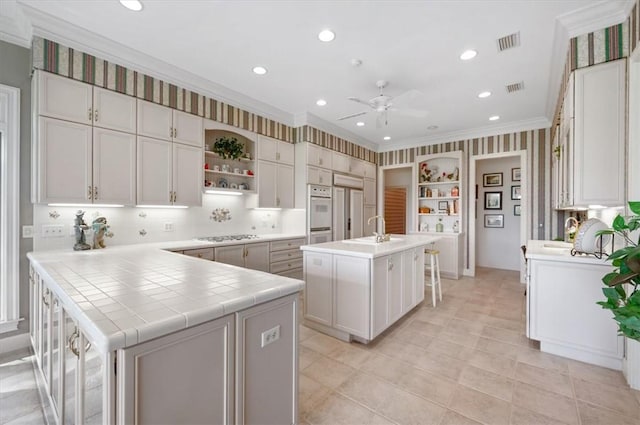 kitchen featuring a kitchen island with sink, white cabinets, crown molding, tile counters, and a breakfast bar area