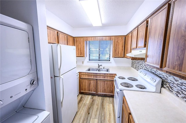 kitchen featuring white appliances, brown cabinetry, under cabinet range hood, a sink, and stacked washing maching and dryer