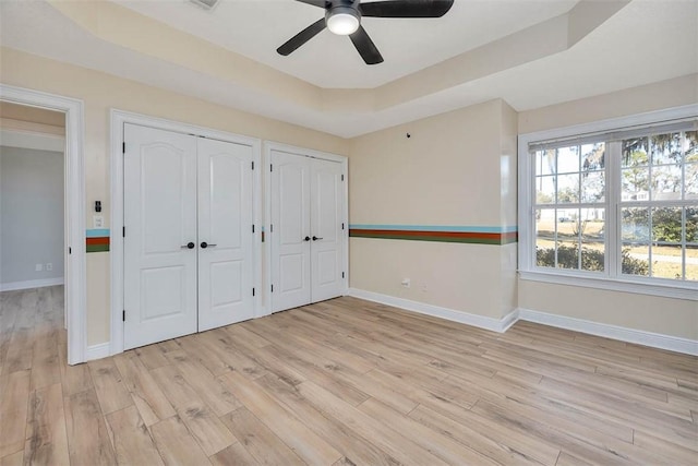 unfurnished bedroom featuring a tray ceiling, light wood-type flooring, two closets, and baseboards