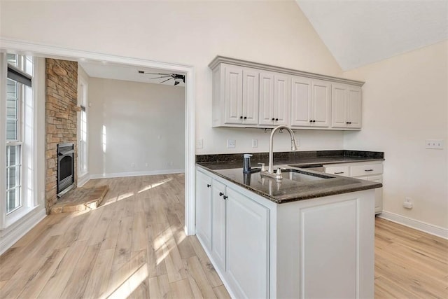 kitchen featuring light wood-style flooring, a ceiling fan, a sink, a stone fireplace, and dark stone counters