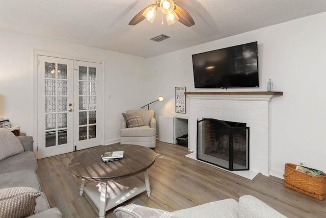 living room with french doors, light hardwood / wood-style flooring, ceiling fan, a textured ceiling, and a fireplace