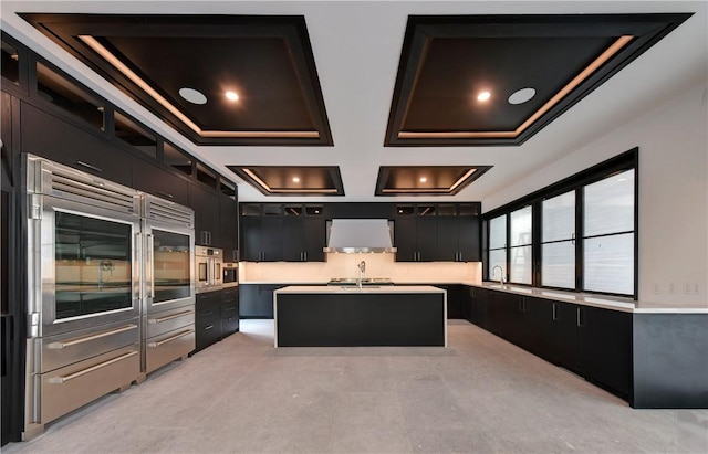 kitchen featuring coffered ceiling, wall chimney range hood, sink, oven, and an island with sink