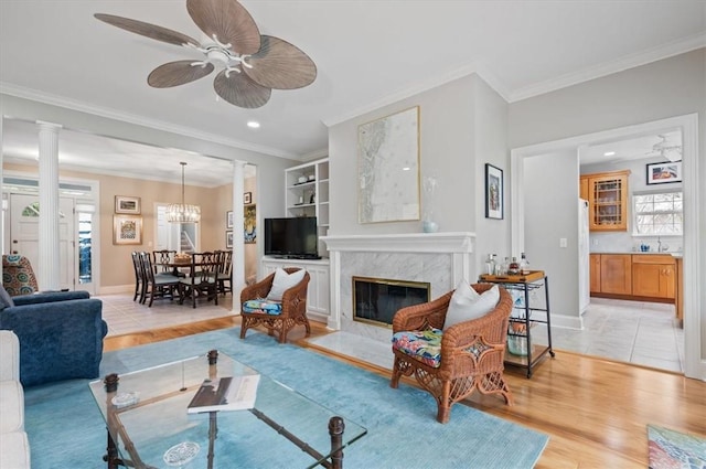 living room with ceiling fan with notable chandelier, a premium fireplace, ornamental molding, ornate columns, and light hardwood / wood-style floors