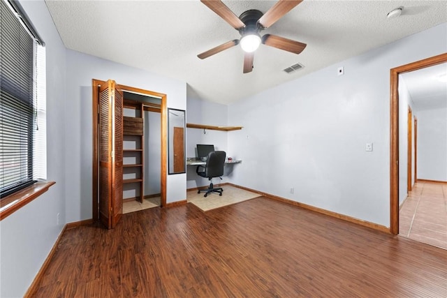 unfurnished office featuring ceiling fan, built in desk, wood-type flooring, and a textured ceiling