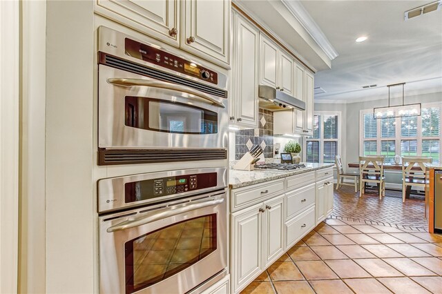 kitchen with crown molding, light tile patterned floors, a notable chandelier, light stone counters, and stainless steel appliances