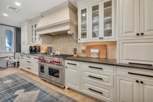 kitchen with premium range hood, backsplash, dark stone counters, double oven range, and white cabinets