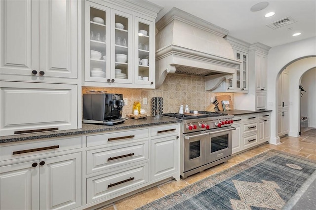 kitchen with white cabinetry, dark stone counters, range with two ovens, decorative backsplash, and custom range hood