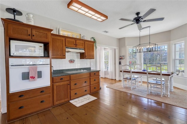 kitchen featuring under cabinet range hood, white appliances, dark wood-style floors, and brown cabinetry