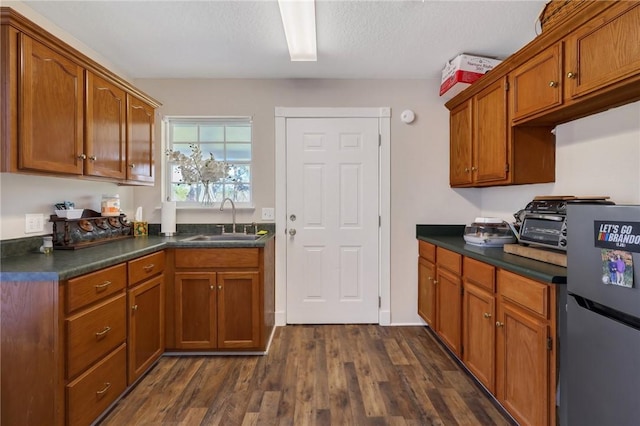 kitchen featuring brown cabinetry, dark wood-style flooring, freestanding refrigerator, a sink, and dark countertops