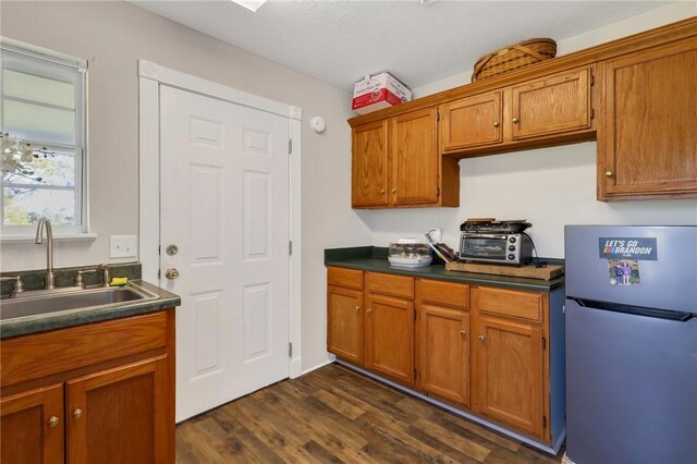 kitchen with a sink, dark countertops, freestanding refrigerator, brown cabinetry, and dark wood-style flooring