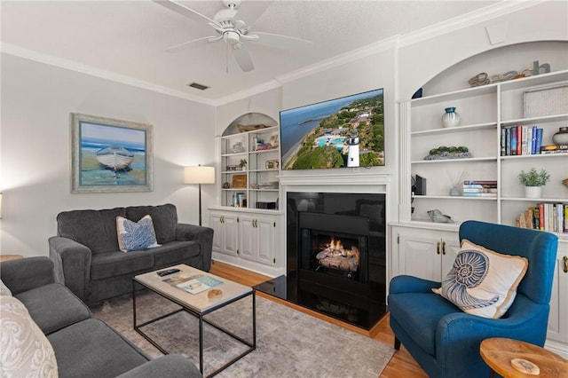 living room with visible vents, ornamental molding, ceiling fan, built in shelves, and light wood-style flooring
