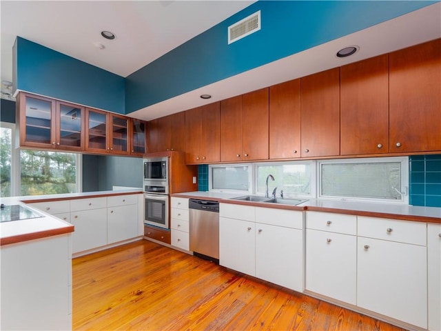 kitchen with a sink, appliances with stainless steel finishes, glass insert cabinets, visible vents, and light wood-style floors