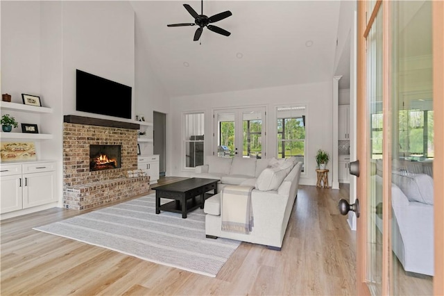 living area featuring high vaulted ceiling, a brick fireplace, a ceiling fan, light wood-style flooring, and french doors