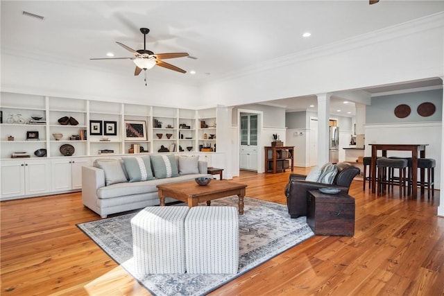 living room with crown molding, built in features, ceiling fan, ornate columns, and light wood-type flooring