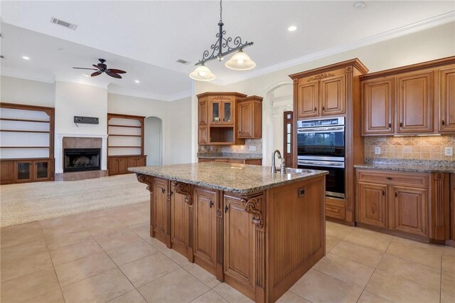 kitchen with crown molding, double oven, hanging light fixtures, light stone countertops, and a center island with sink