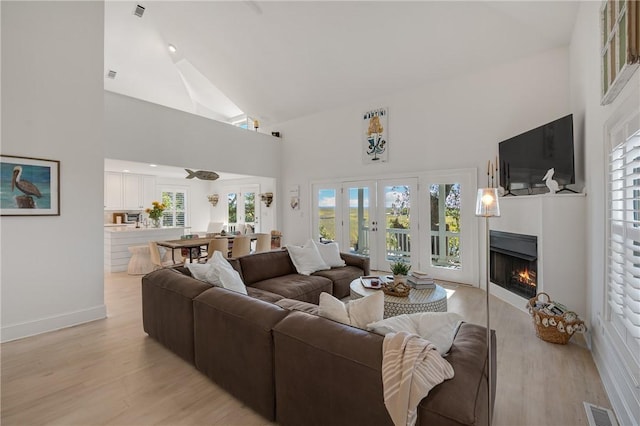 living room featuring a warm lit fireplace, high vaulted ceiling, light wood-type flooring, and visible vents