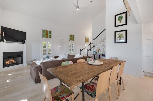 dining space with stairway, a towering ceiling, ceiling fan, light wood-type flooring, and a warm lit fireplace