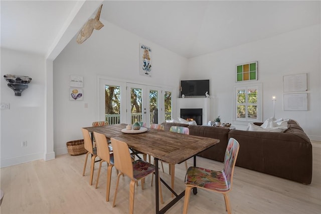 dining room featuring light wood-type flooring, a lit fireplace, high vaulted ceiling, and baseboards