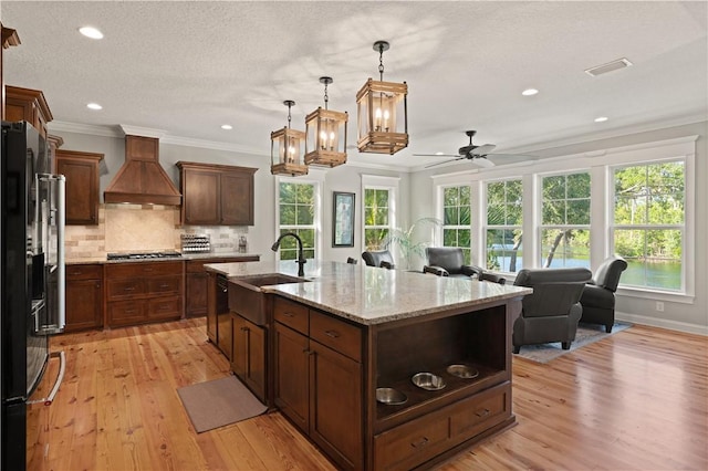 kitchen featuring stainless steel gas stovetop, sink, an island with sink, custom range hood, and fridge with ice dispenser