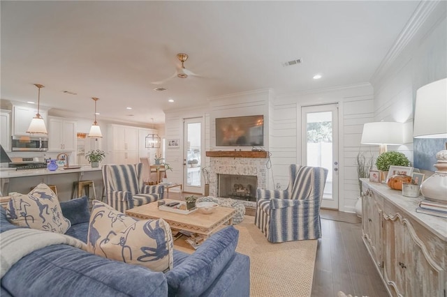 living room featuring ceiling fan, a stone fireplace, crown molding, and dark wood-type flooring