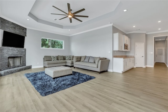 living room with ceiling fan, a raised ceiling, a stone fireplace, light wood-type flooring, and ornamental molding