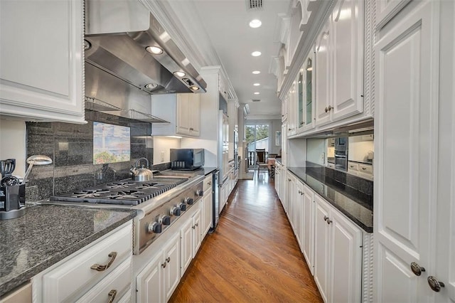 kitchen with wall chimney exhaust hood, dark stone countertops, hardwood / wood-style floors, decorative backsplash, and white cabinets