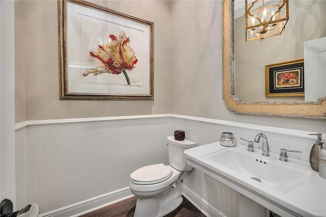 half bathroom featuring toilet, a notable chandelier, wood finished floors, a sink, and wainscoting