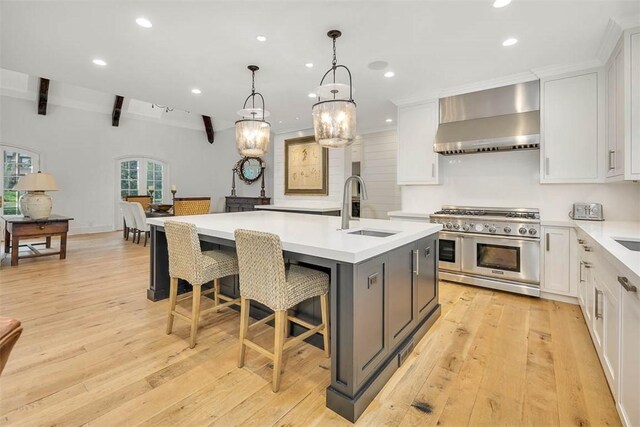 kitchen featuring double oven range, a center island with sink, wall chimney range hood, light wood-type flooring, and beam ceiling