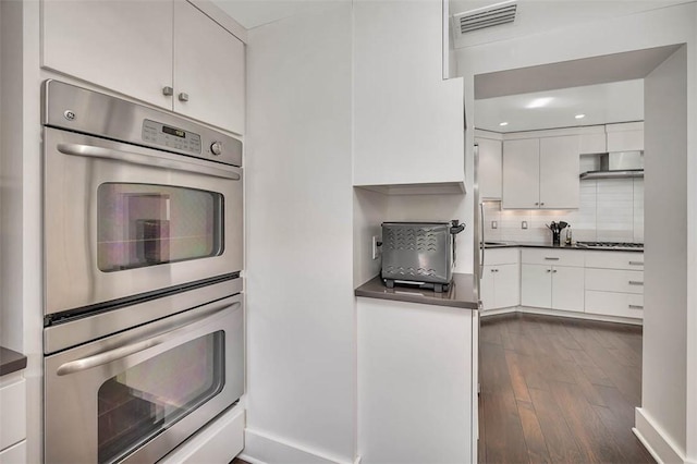 kitchen with tasteful backsplash, white cabinets, dark countertops, wall chimney exhaust hood, and stainless steel double oven