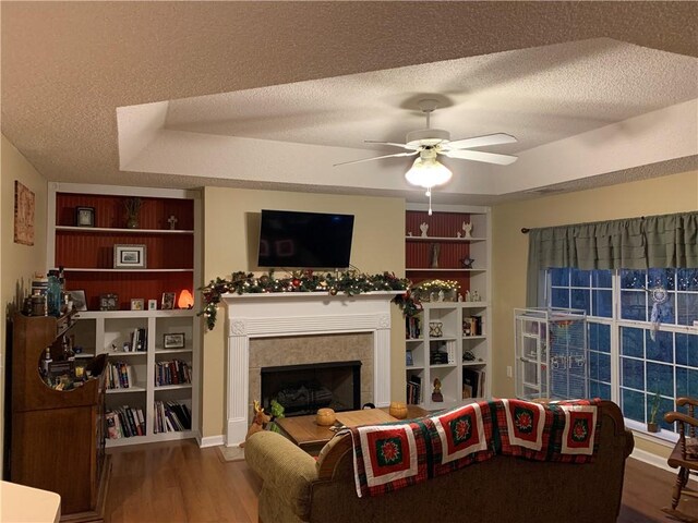 living room featuring built in shelves, a textured ceiling, wood finished floors, a raised ceiling, and a tile fireplace