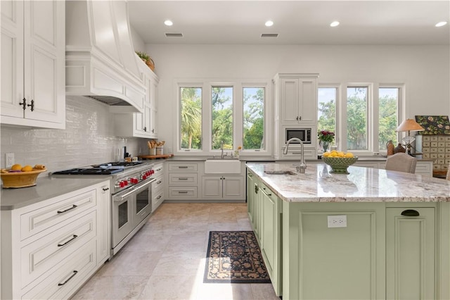 kitchen with a wealth of natural light, stainless steel appliances, green cabinets, premium range hood, and an island with sink