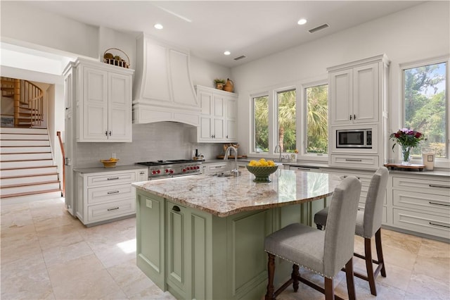 kitchen featuring sink, light stone countertops, an island with sink, appliances with stainless steel finishes, and custom range hood