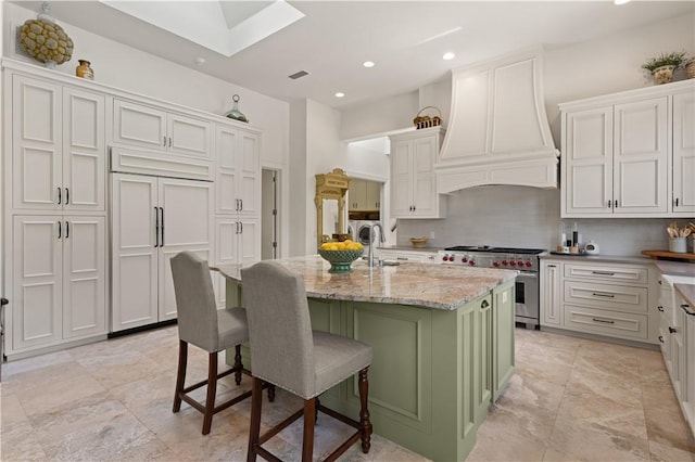 kitchen featuring a kitchen island with sink, high end stainless steel range, light stone countertops, a kitchen bar, and custom range hood