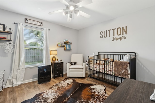 bedroom with wood finished floors, ceiling fan, a nursery area, and baseboards