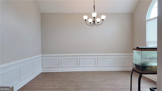 unfurnished dining area with an inviting chandelier, a decorative wall, light wood-type flooring, wainscoting, and vaulted ceiling