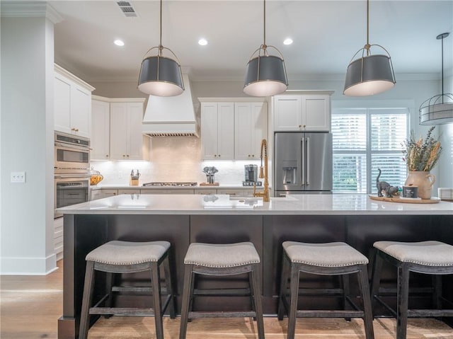 kitchen featuring an island with sink, stainless steel appliances, decorative light fixtures, light wood-type flooring, and white cabinets