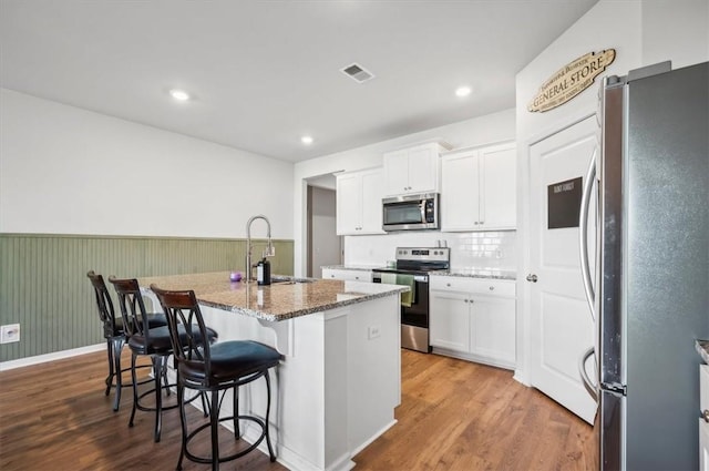 kitchen with stainless steel appliances, an island with sink, stone countertops, white cabinets, and hardwood / wood-style flooring